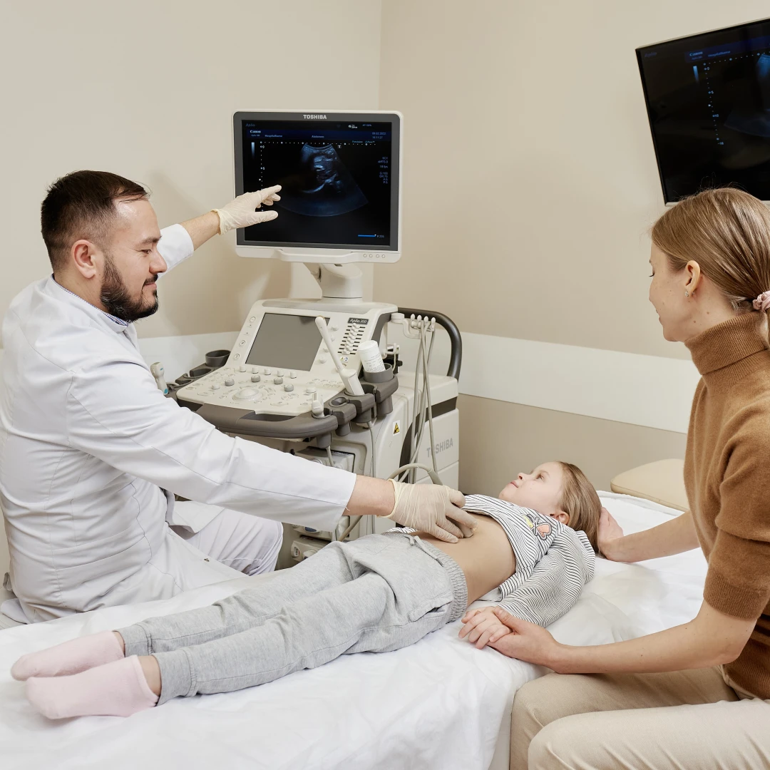 Мужчина врач в белом халате делает УЗИ желчных протоков девочке лет 10, лежащей на кушетке. Рядом сидит мама и держит девочку за руку.