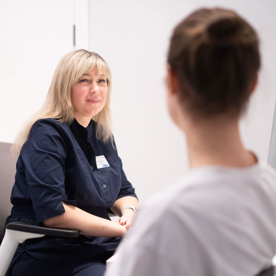 Женщина врач в темно-синем медицинском костюме сидит за рабочим столом и выслушивает пациентку, сидящую спиной к камере.