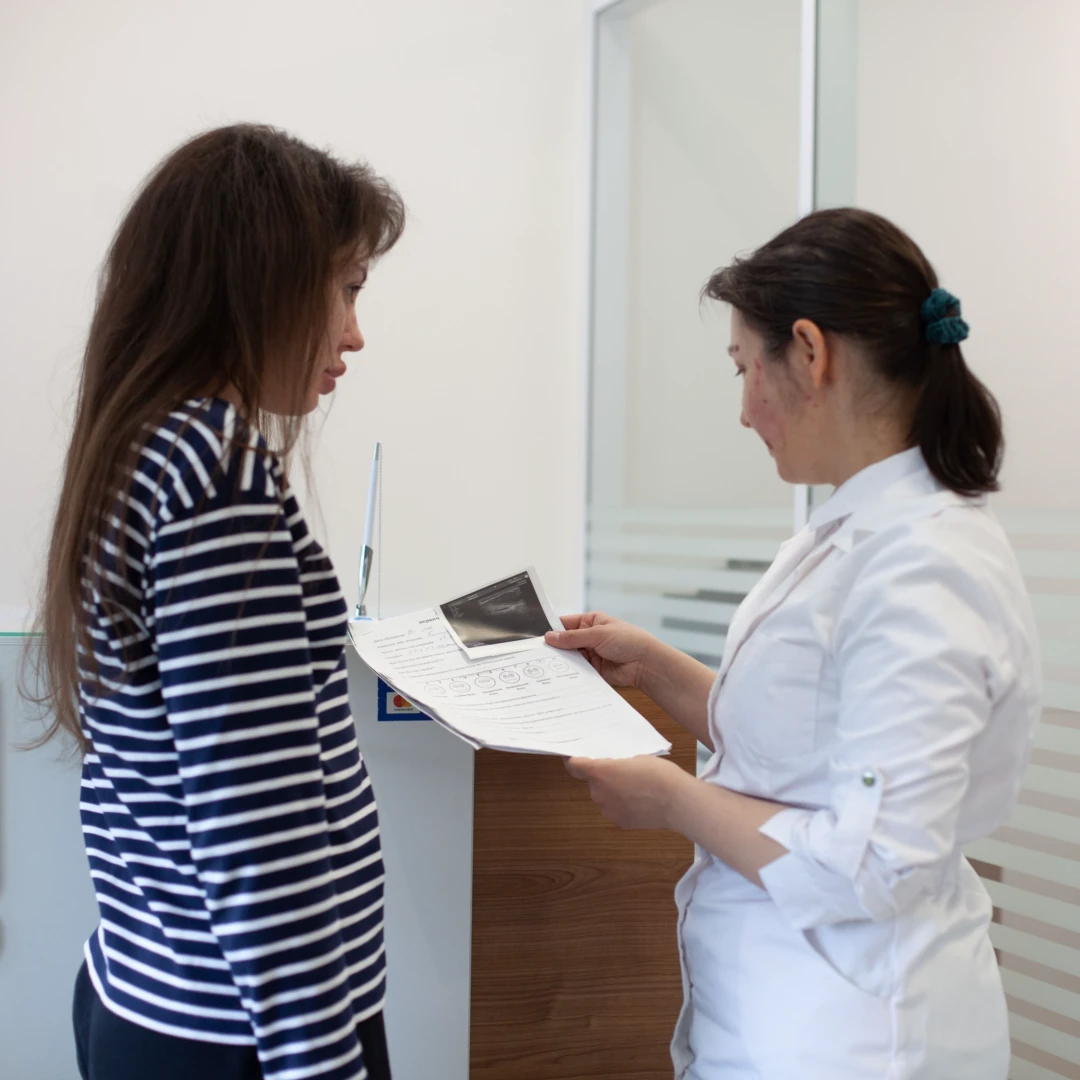 Женщина врач в белом халате дает пациентке разъяснения после УЗИ желчных протоков, держа в руках заключение и снимки УЗИ.