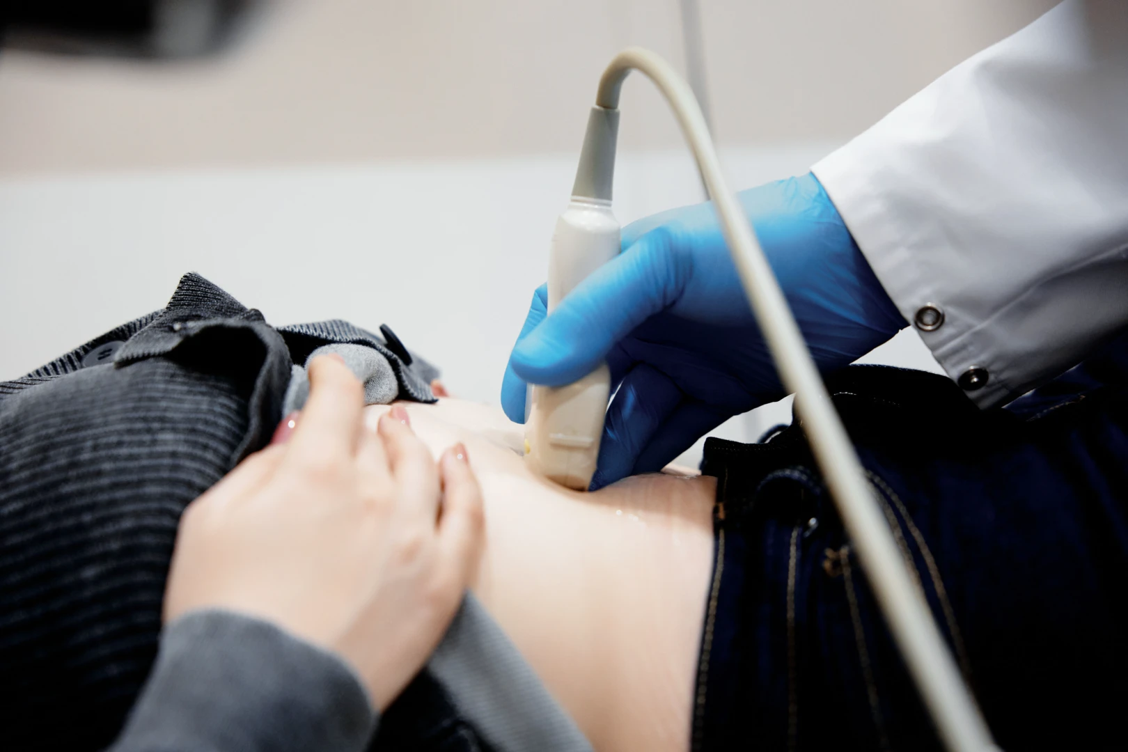 Женщина врач в белом халате проводит УЗИ мягких тканей с эластометрией пациенту, лежащему на кушетке.