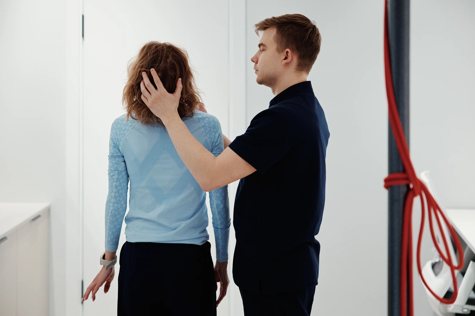 Специалист осматривает осанку стоящей пациентки в кабинете, придерживая её за шею и плечо
