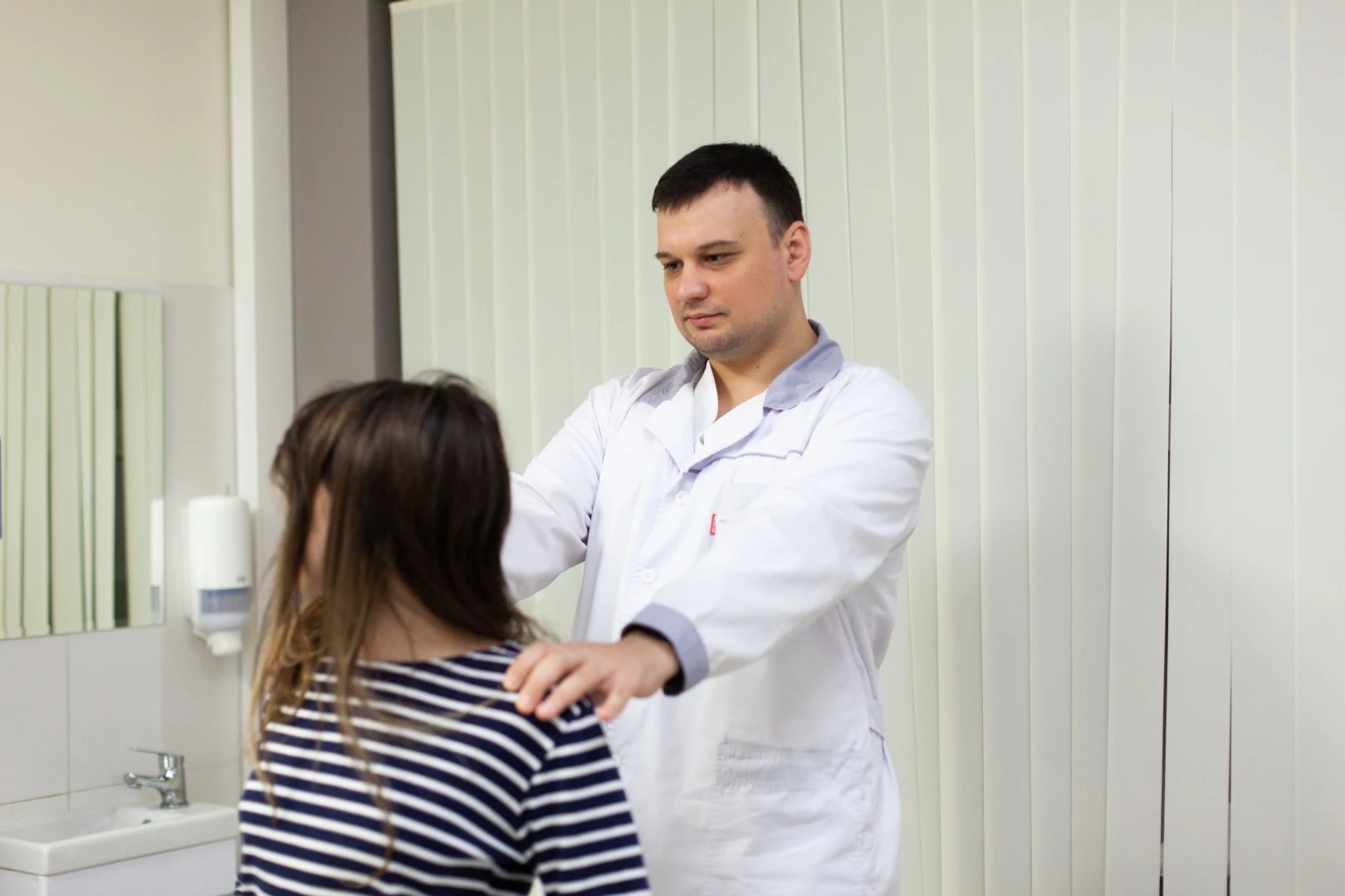 Врач сравнивает положение плеч пациентки, стоя позади неё
